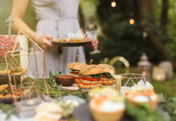 Young lady standing behind a table holding a plate. The table has several food items like burgers.