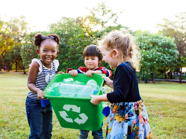 Three children holding a green recycling bin full of plastic.